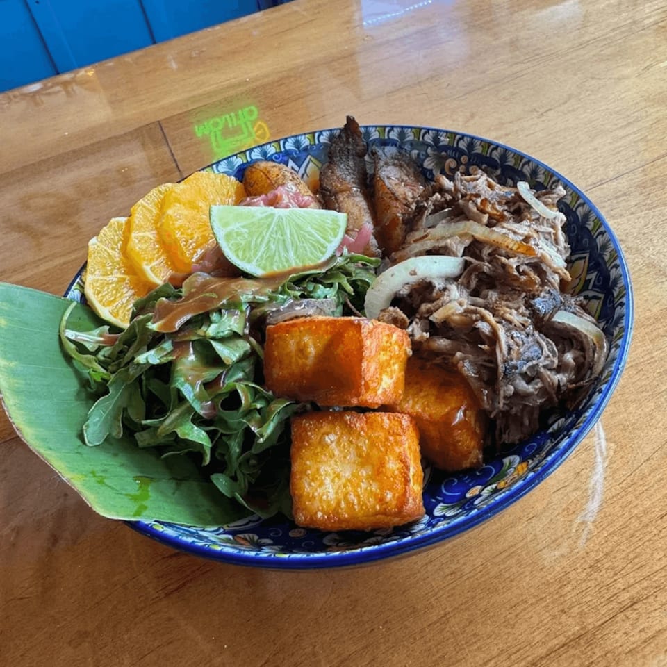 A vibrant Cuban dish with shredded meat, fried plantains, salad, orange slices, and a lime wedge.