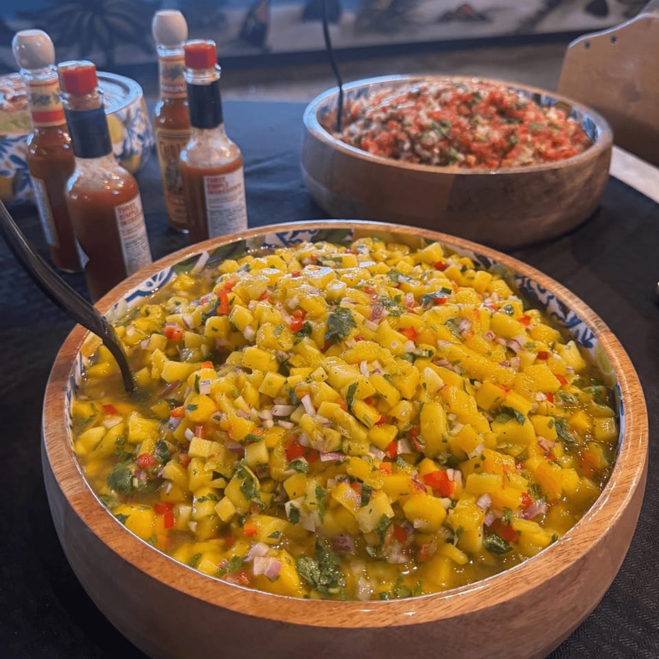 A large wooden bowl of vibrant mango salsa with red onion and cilantro, alongside hot sauce bottles.