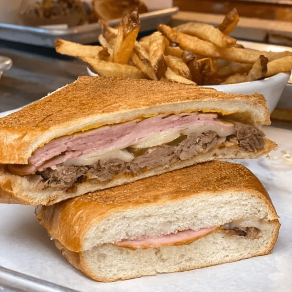 A sliced Cuban sandwich with ham, pork, cheese, and pickles, served with a side of fries.