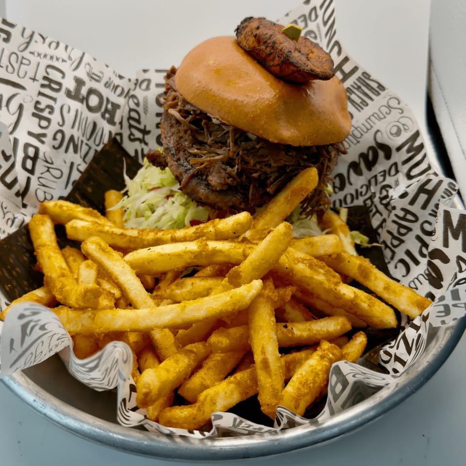A Cuban pulled pork sandwich with plantain on top, served with a side of seasoned fries.