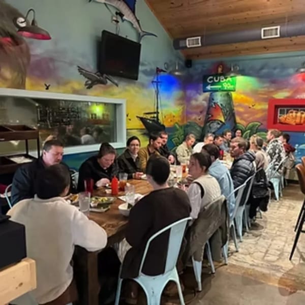 Diners seated at a long table in The Hush Cuban Kitchen, with a vibrant mural in the background.