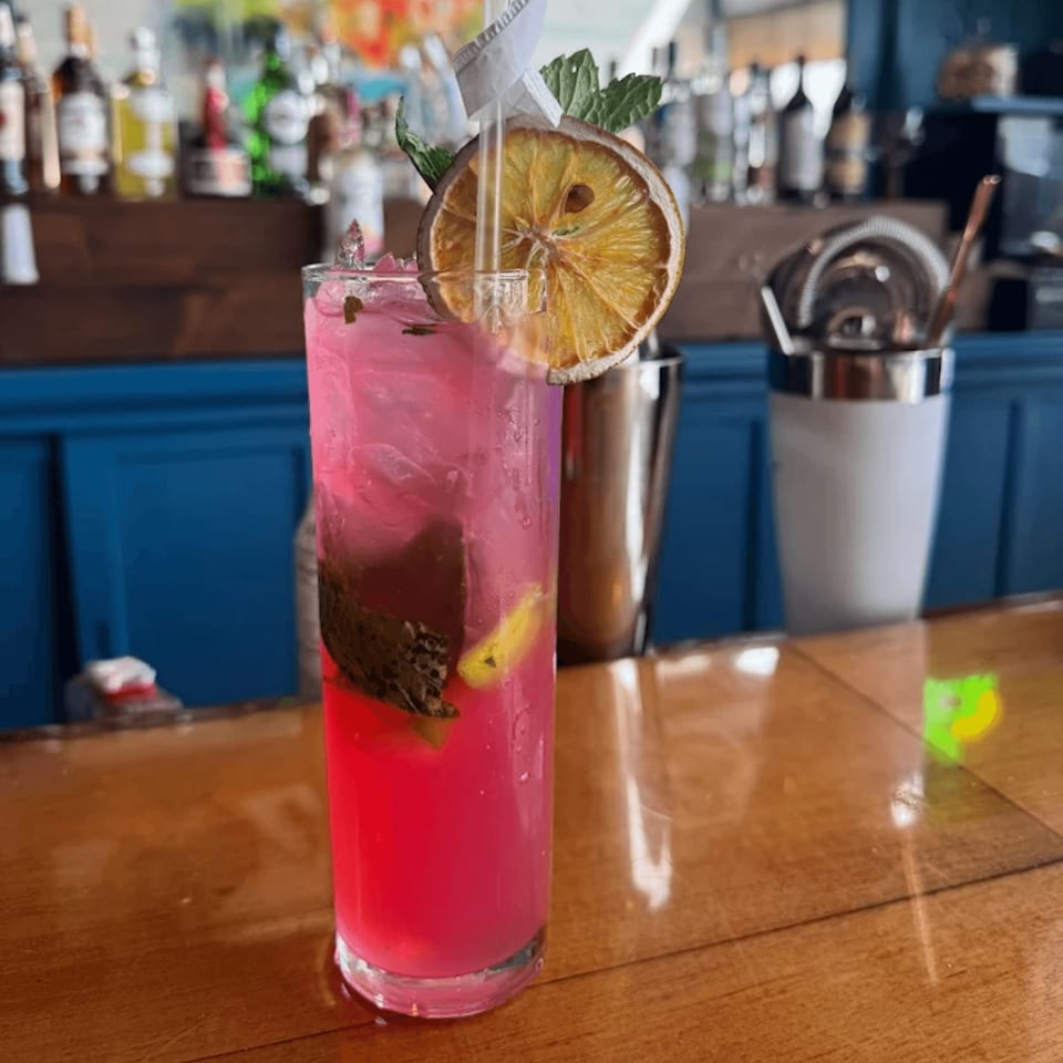 A vibrant pink Cuban cocktail with mint, dried orange, and a straw on a wooden bar.