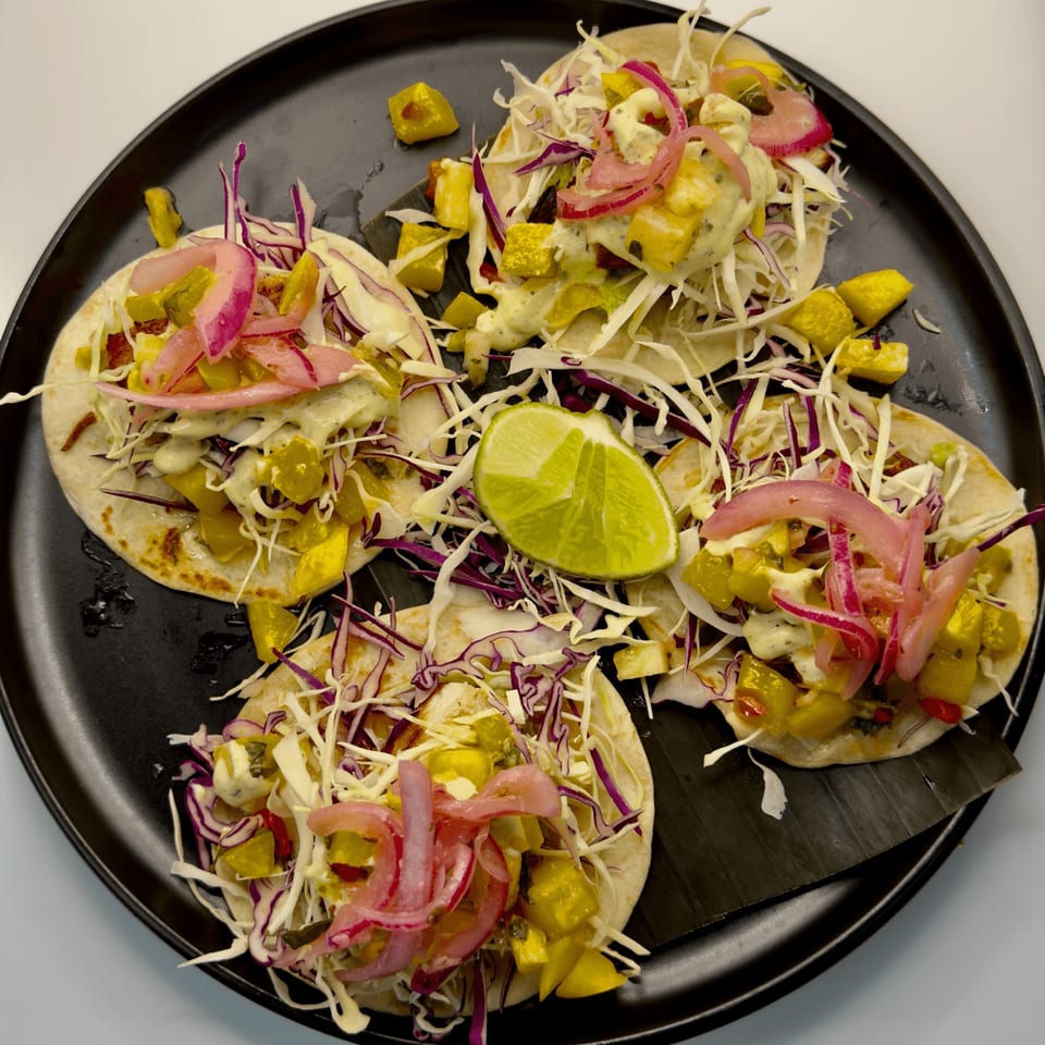 Four Cuban tacos with plantains, pickled red onions, cabbage, and a lime wedge on a black plate.