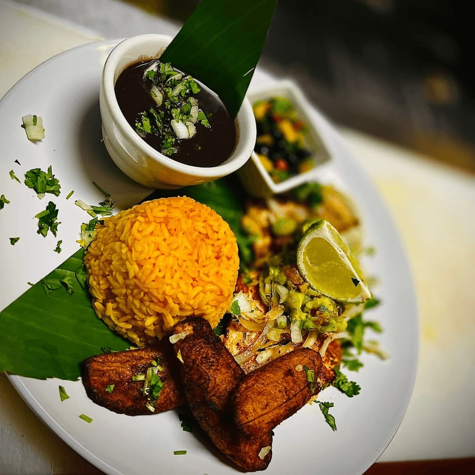 Cuban dish with yellow rice, fried plantains, black bean soup, and fish with salsa on a white plate.