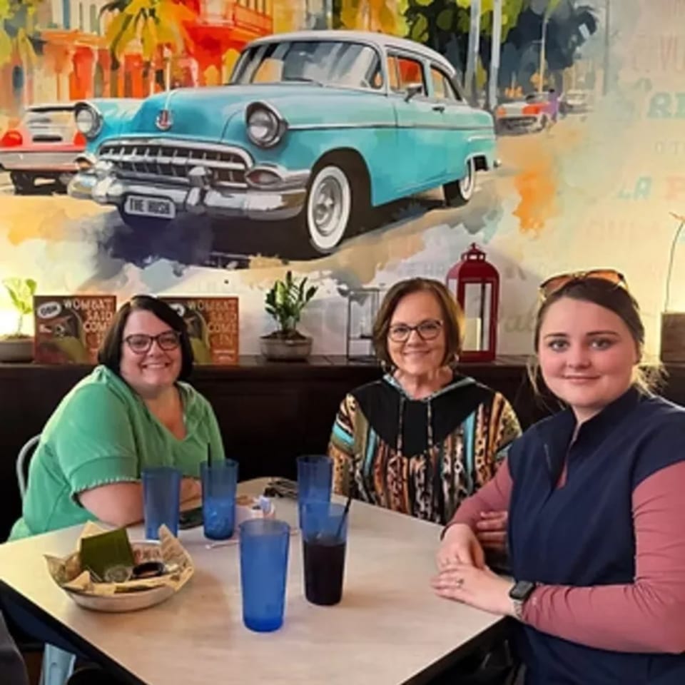 Three women smiling at a table in The Hush Cuban Kitchen, with a vibrant Cuban car mural behind them.