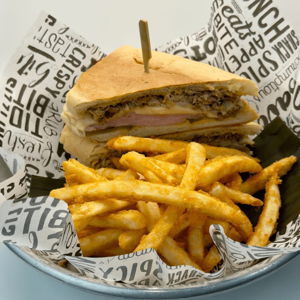 A Cuban sandwich with a toothpick, served with a side of golden french fries in a metal basket.