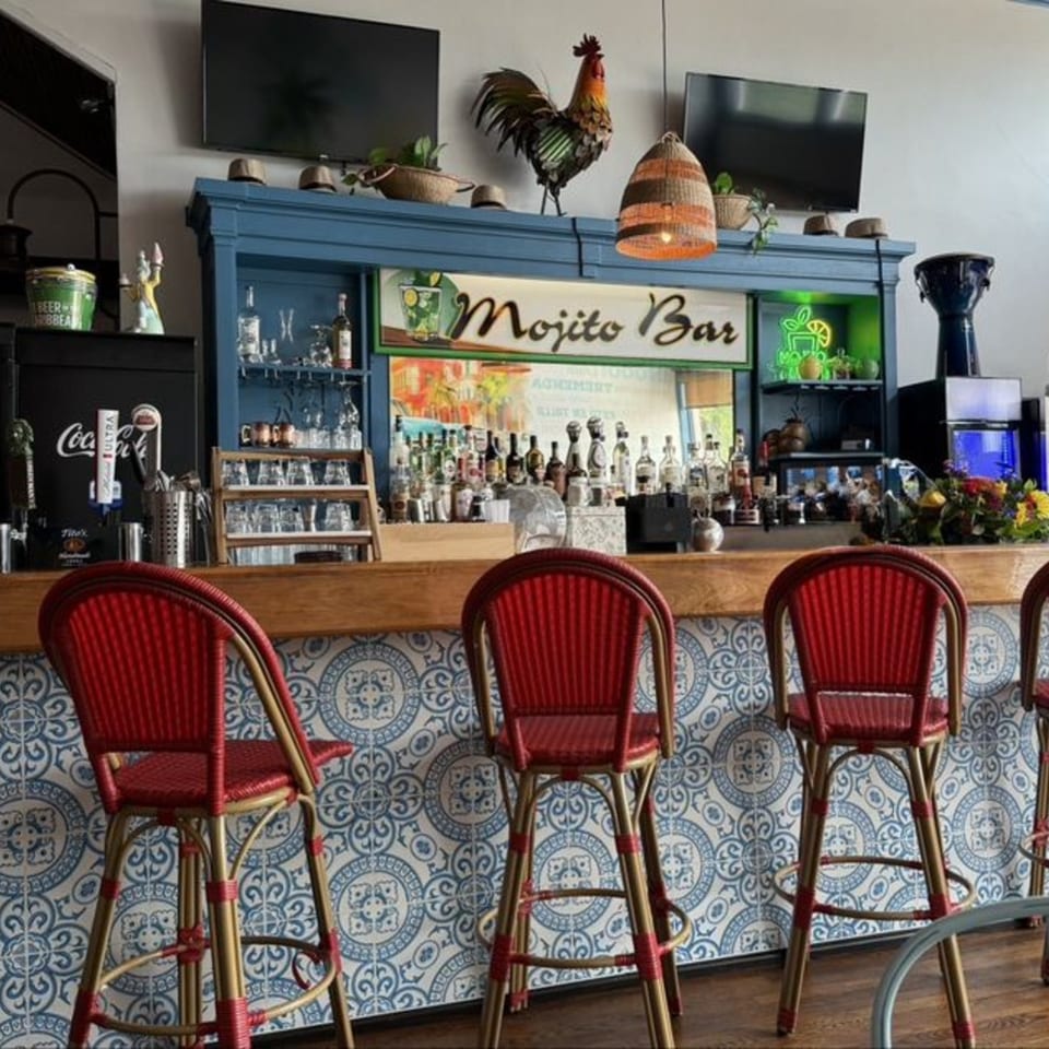 A vibrant Cuban Mojito Bar with red stools, blue tiled counter, and a rooster statue.